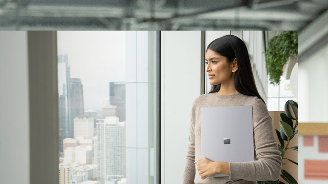 Seseorang memegang peranti Surface Laptop 5 pada badan mereka dengan satu tangan sementara melihat ke seberang bilik