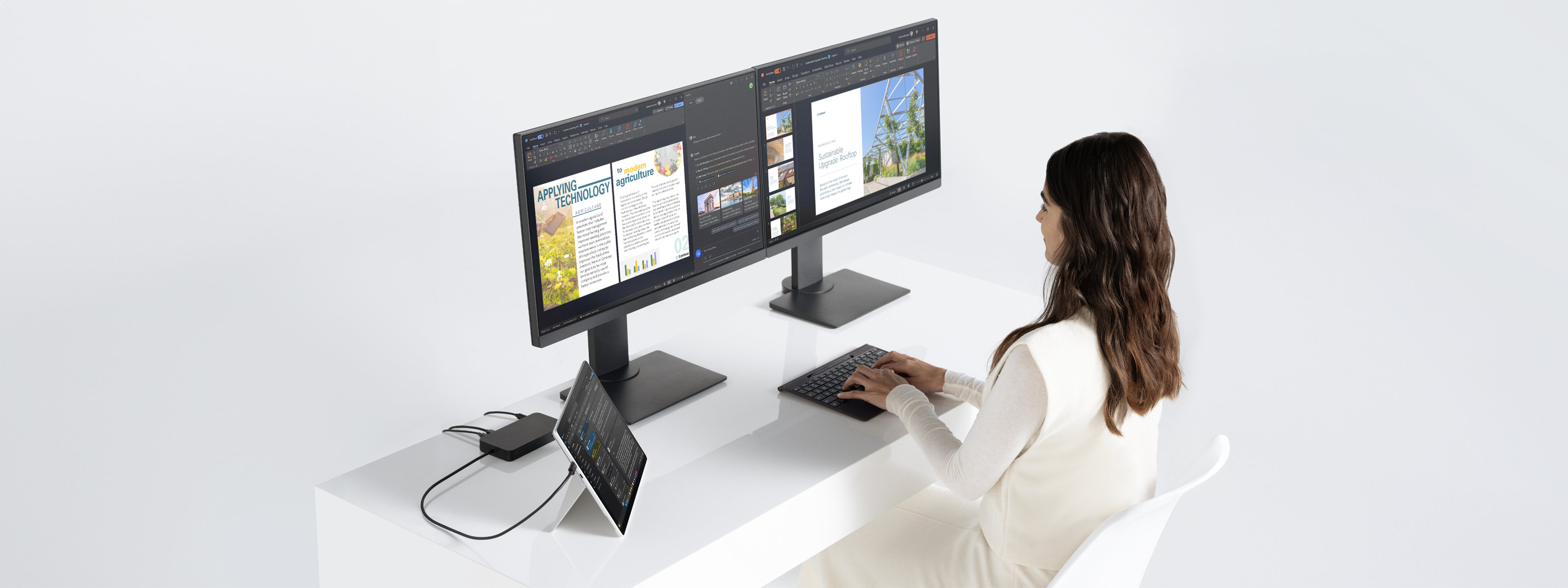 A woman sits at a desk with a Surface Pro device in kickstand mode, connected to two monitors through a dock while she works.