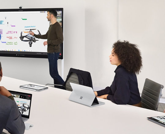 Dos trabajadores colaboran mediante Surface Hub 2S mientras lideran una reunión virtual en Teams en un dispositivo Surface Laptop sobre un escritorio cercano