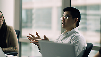 En mand med briller gestikulerer, mens han taler ved siden af sin bærbare computer i et kontormiljø