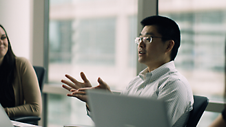 A man wearing glasses gesturing while speaking next to his laptop in an office environment