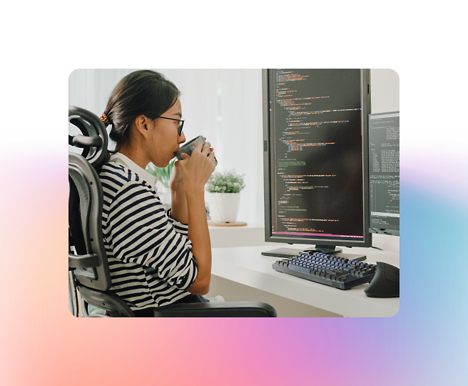 A person sits at a desk sipping from a mug while looking at code on a computer screen