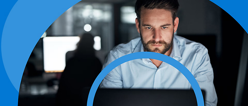 Man with a beard working intently on a laptop in a modern office environment during evening hours.