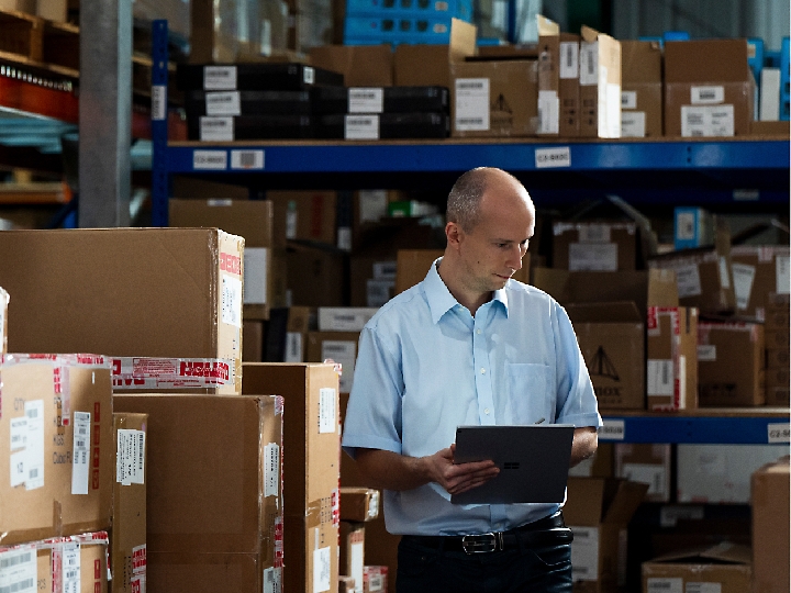 A man in a light blue shirt stands in a warehouse filled with cardboard boxes, holding a tablet.