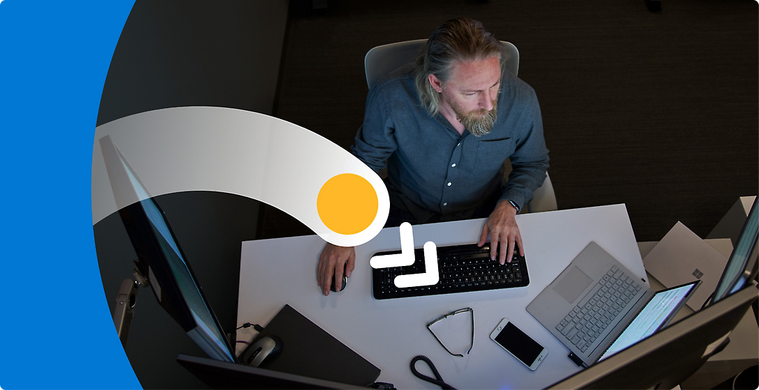 A man with a beard working at a desk in a dimly lit office, focusing on computer screens.