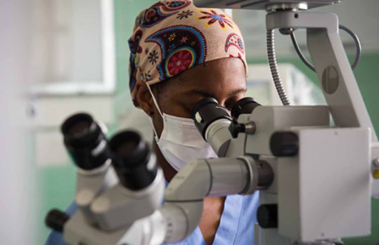 Una persona con una mascarilla y un pañuelo en la cabeza mira a través de un microscopio.