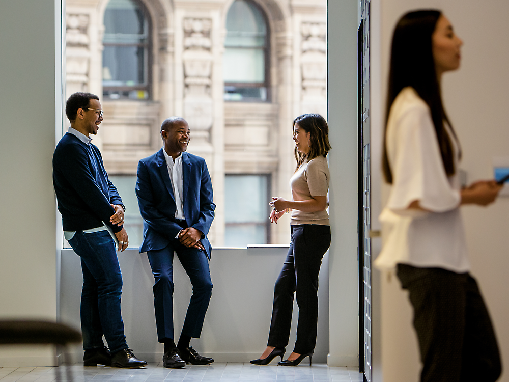 Four colleagues conversing casually in an office, with a cityscape visible through the window.