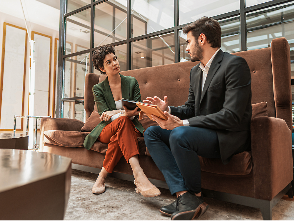 Two professionals sitting on a sofa and engaged in a serious conversation, one holding a digital tablet.