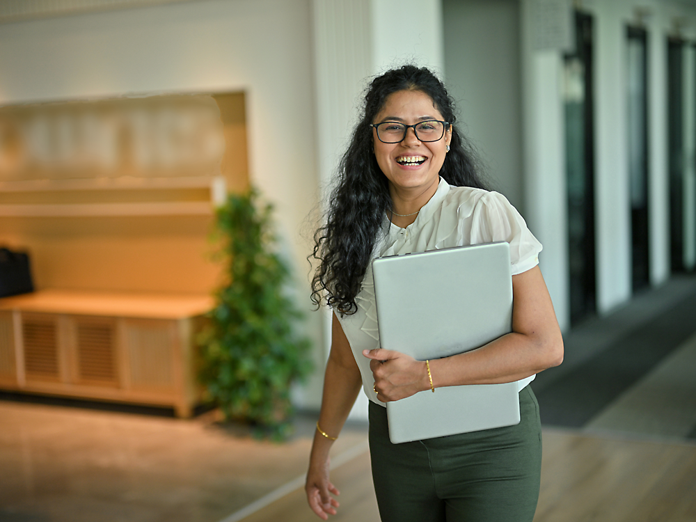 A cheerful woman with curly hair, wearing glasses and office attire, holds a laptop while standing in an office hallway.