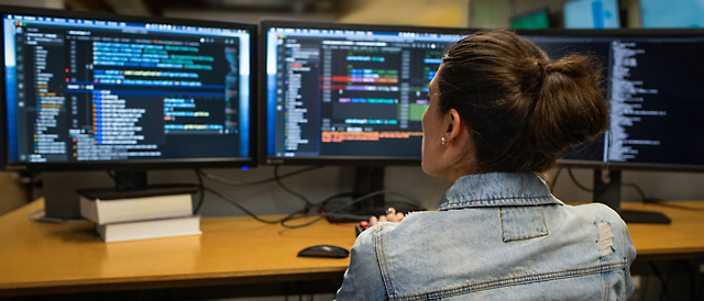 Kvinne som programmerer ved hjelp av flere skjermer som viser kode i et mørkt kontorlandskap.