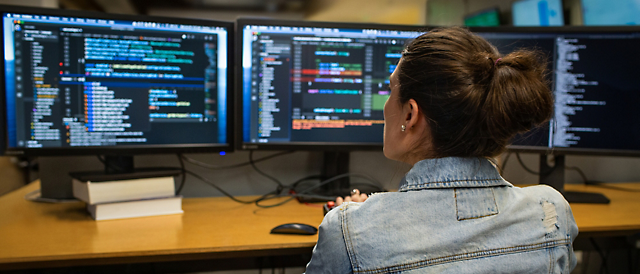 Vrouw die codeert op meerdere computerschermen waarop softwarecode wordt weergegeven in een zwak verlicht kantoor.