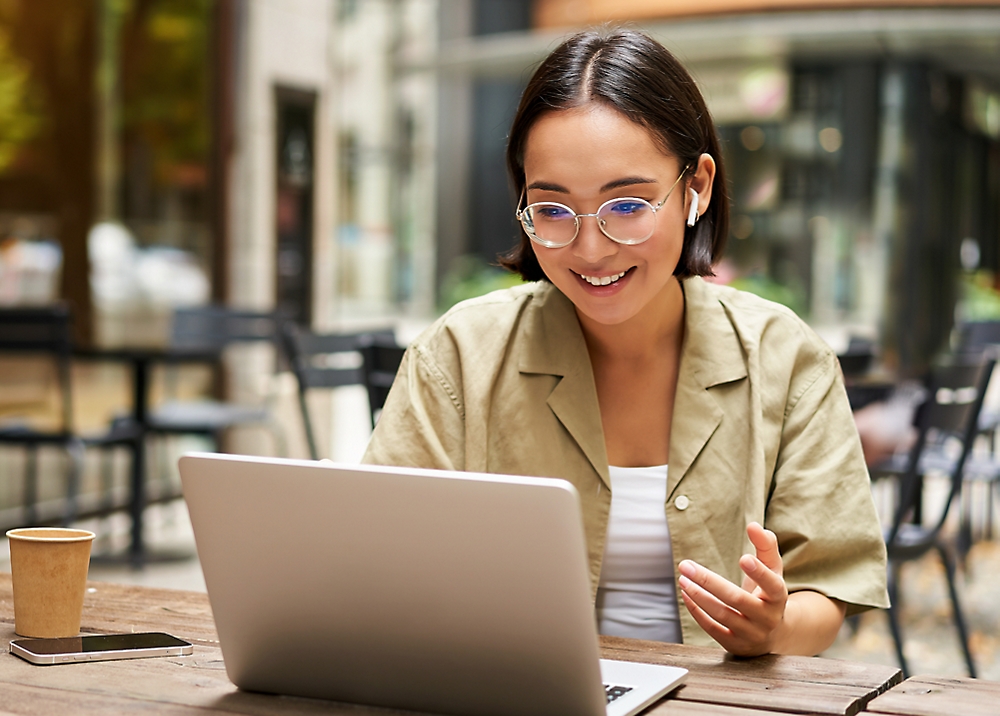 Young woman wearing glasses and smiling while using a laptop at an outdoor cafe table, with a smartphone