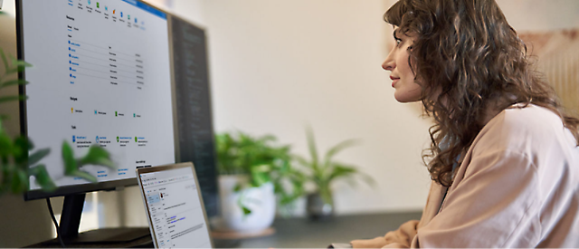 Vrouw die aan een bureau werkt met een computermonitor die een spreadsheet en een laptop aan de zijkant weergeeft