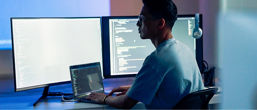 A person is sitting at a desk in front of multiple computer screens displaying code, working on a laptop in a dimly lit room.