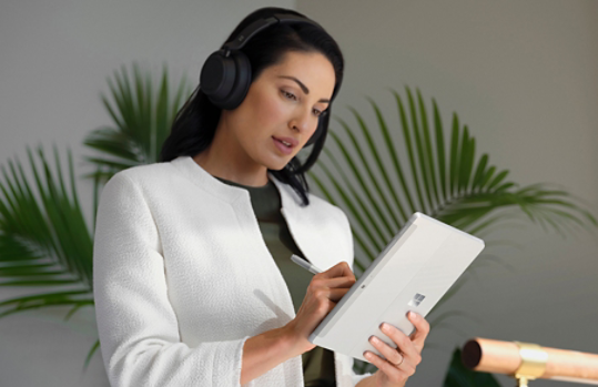 Seseorang yang memakai Surface Headphones 2+ menggunakan Pen Surface untuk menavigasi Surface Go 4 dalam mod tablet di tempat kerja