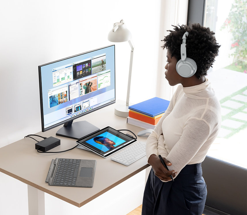 Osoba nosząca słuchawki Surface Headphones, stojąca przed biurkiem, na którym znajduje się urządzenie Surface, klawiatura Type Cover i monitor.