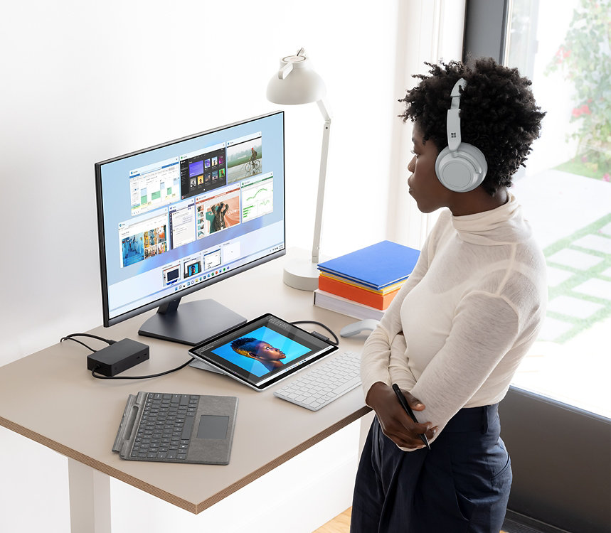 A person wearing Surface Headphones standing in front of a desk with a Surface device, Type Cover, and monitor on it.