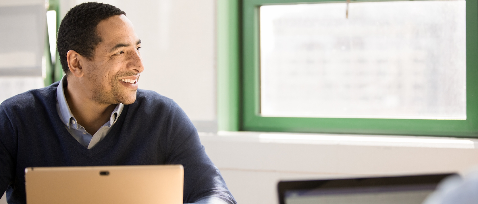 A man is smiling at a laptop in front of a window.