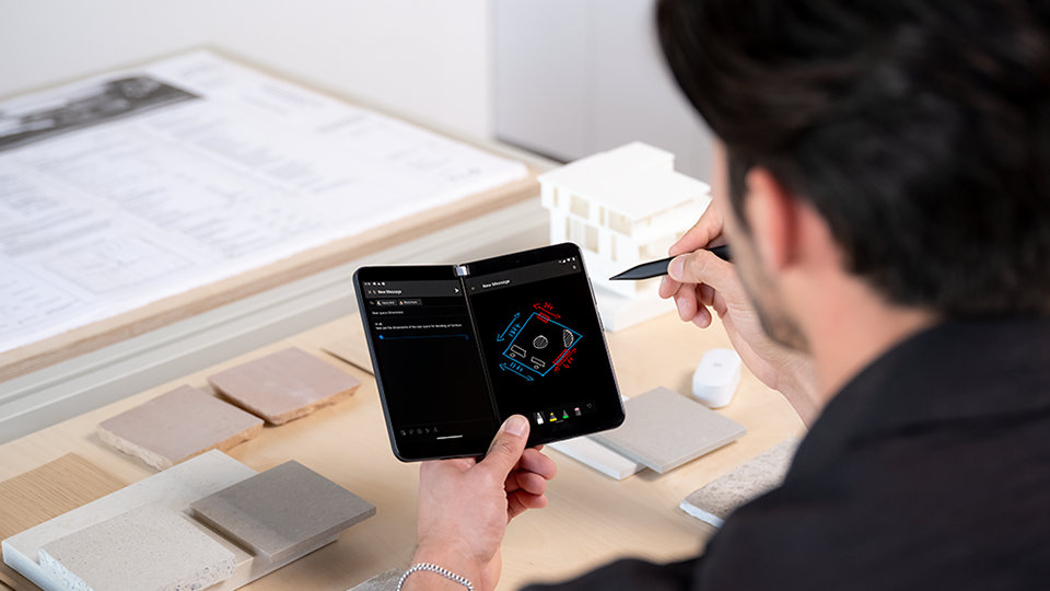 A person using a Surface Slim Pen on a Surface Duo at a desk with blueprints and stone samples..