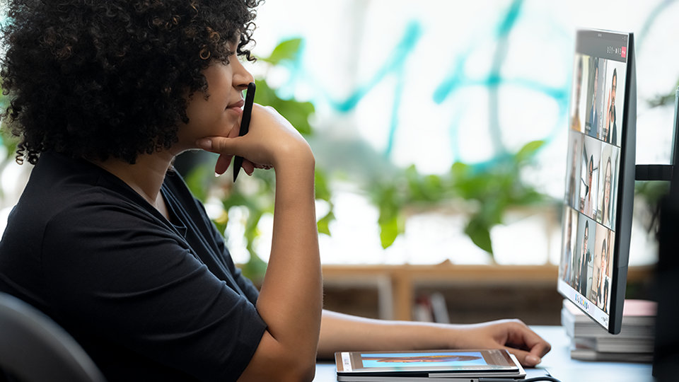 A person sitting at a desk holding a Slim Pen with a Surface Laptop Studio in tablet position flat on desk and a Microsoft Teams meeting on a monitor.