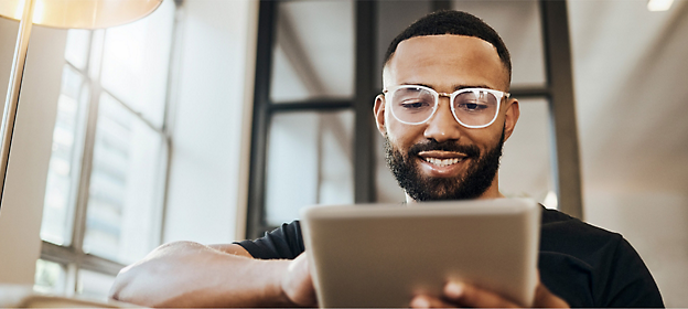 Un homme porte des lunettes, un tee-shirt noir, tient une tablette et sourit.