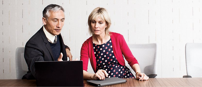 Um homem e uma mulher sentados em uma mesa de conferência olhando para um laptop.