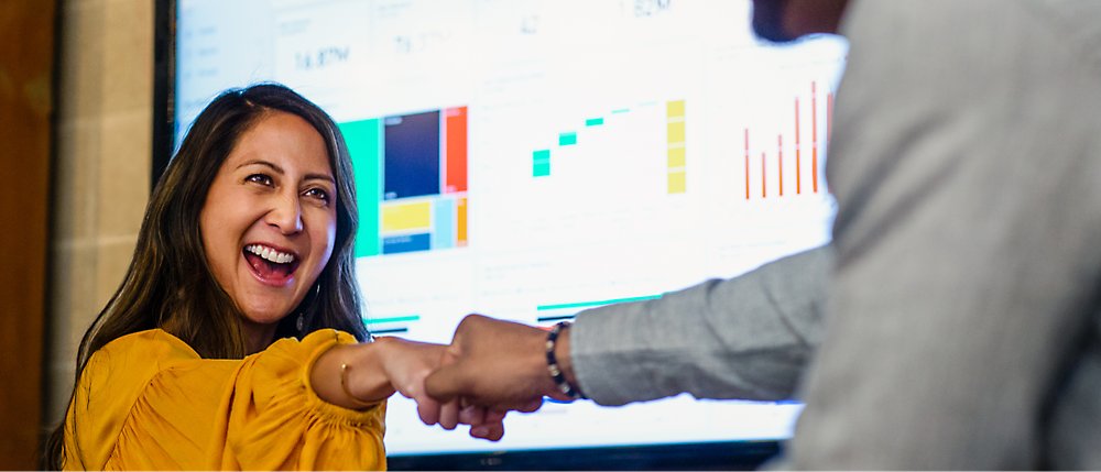 Egy sárga felsőt viselő nő mosolyog és ökölpacsizik egy kollégájával a grafikonokat és diagramokat megjelenítő képernyő előtt.