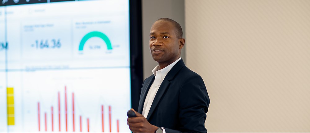 A man dressed in a dark blazer and white shirt stands in front of a large screen displaying various charts and graphs