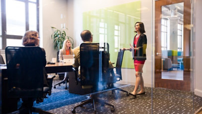 会議室で、立っている女性のビジネス パーソンが、テーブルに着席している複数のビジネス パーソンに話しています。