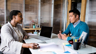 A woman and a man talking in her office with a laptop on the desk.