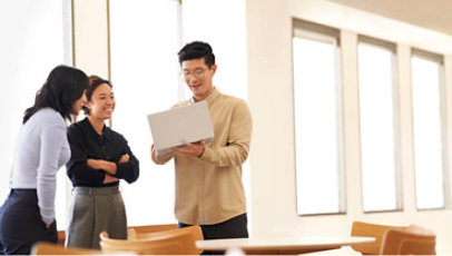 Three employees stand around a laptop computer in a bright office setting.