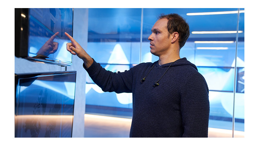 A man wearing a hooded sweatshirt inside a secure office room pointing at a geographic area displayed on a large monitor.