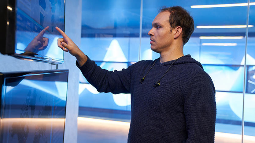 A man wearing a hooded sweatshirt inside a secure office room pointing at a geographic area displayed on a large monitor.