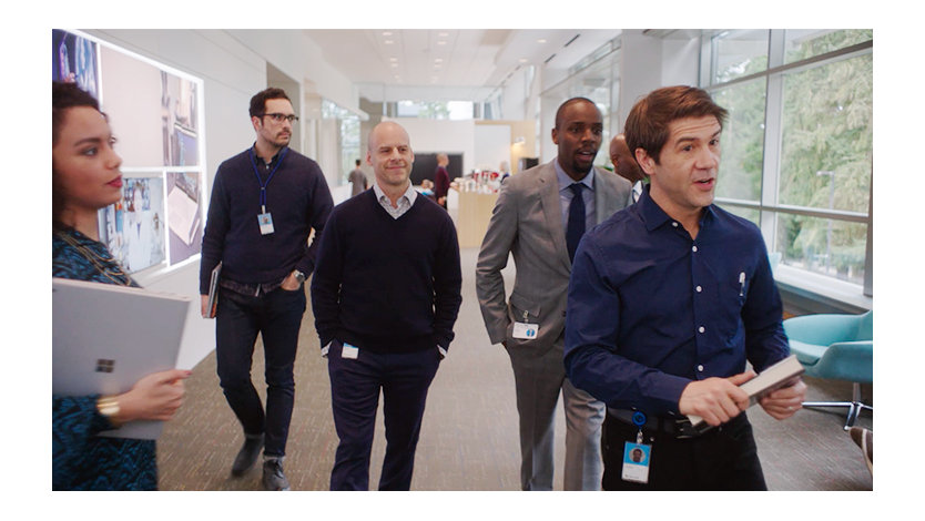 John Thompson from the SBC video is leading a group of five office workers down an office hallway while they carry on a conversation.