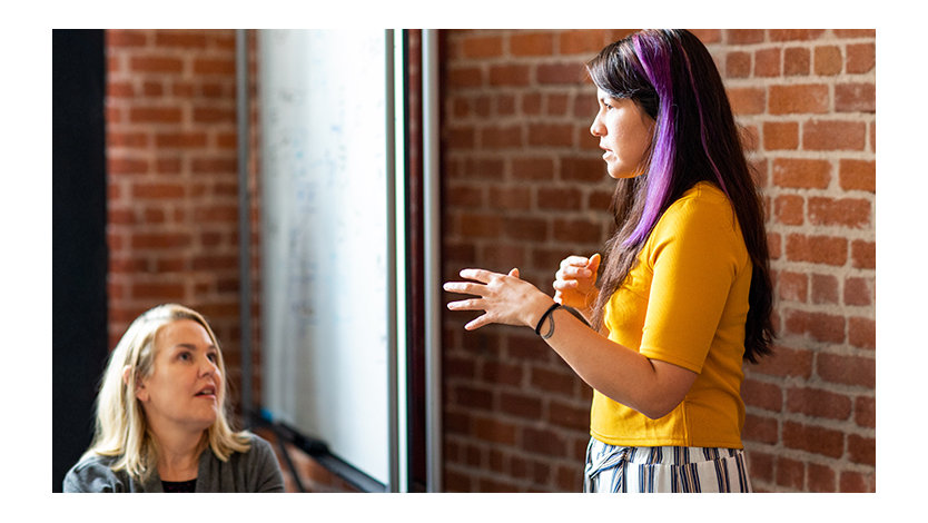 A female developer is speaking near a white board during team stand up meeting.