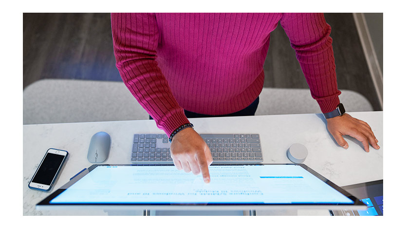 Top-down view of a man wearing a dark red shirt working on a Microsoft Surface Studio with a phone next to him on the desktop along with a mouse, keyboard, and Surface Dial.