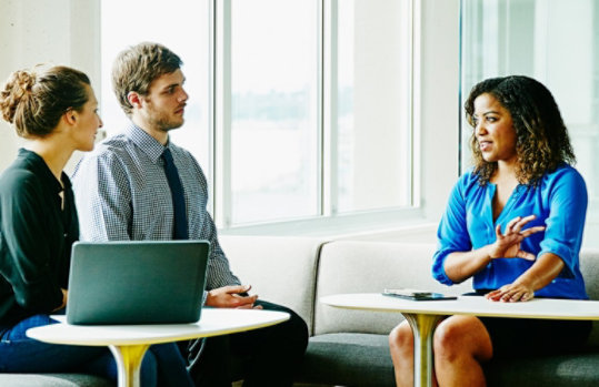 A businesswoman leads a meeting with two colleagues in a sunny office.