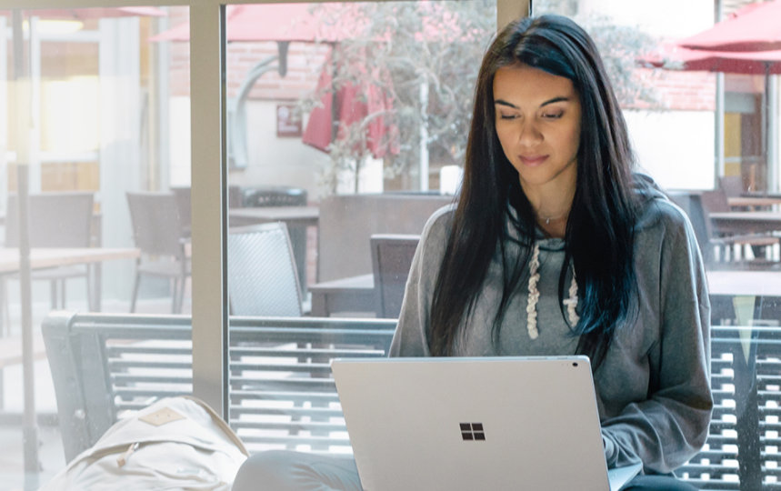 A young woman is sitting cross-legged on a windowsill using a Surface Book on her lap.