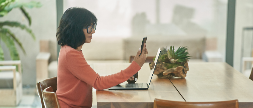 Жена с очила, използваща лаптоп и смартфон на модерна дървена маса, със саксийно растение 