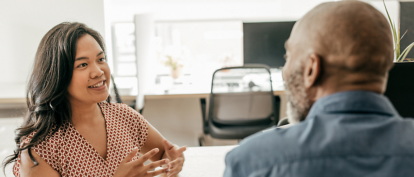 A woman and a man engaged in a conversation at a table in a brightly lit office setting.