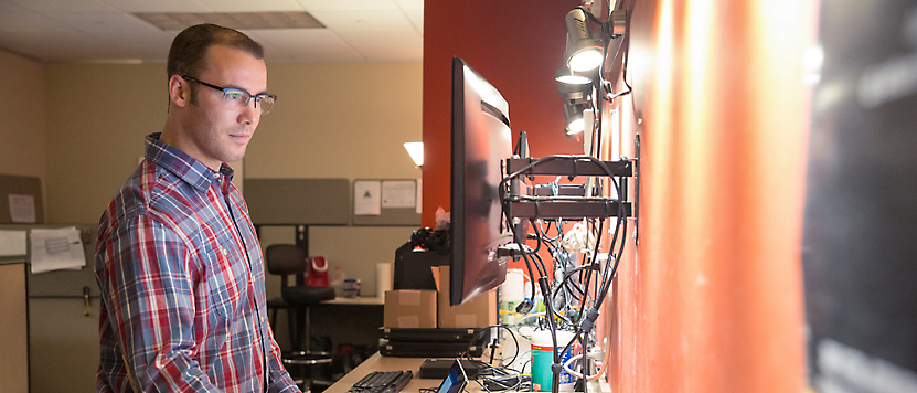 A man wearing glasses and a plaid shirt concentrates on a computer screen in a cluttered office environment.