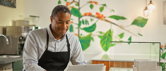 A man in an apron is using a tablet in a coffee shop.