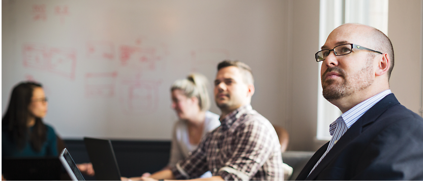 Business professionals in a meeting room with one focused individual in the foreground and a whiteboard with notes in the background.