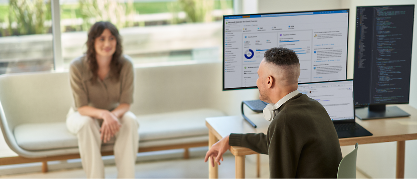 Two professionals in an office setting, with one person seated at a computer workstation and the other seated across, having a discussion.