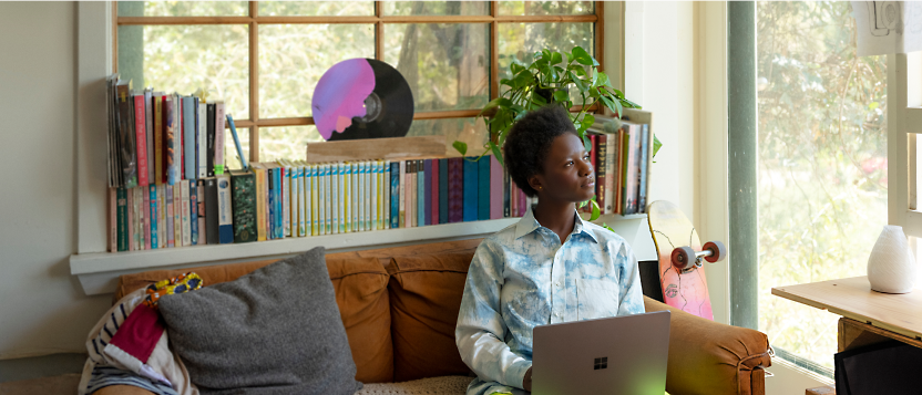 Woman sitting on a couch with a laptop, looking thoughtfully to the side in a cozy room with a bookshelf and skateboard.