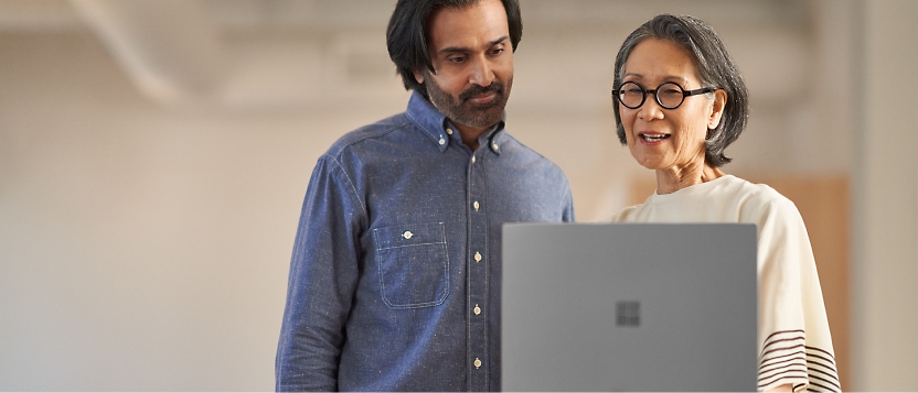 Two people stand side by side, looking at a laptop screen. One is wearing a blue shirt, and the other is wearing glasses.