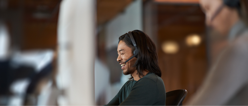 Asiatisk mand med headset smiler under en samtale i et moderne kontormiljø.