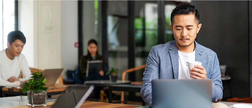 Drie mensen die op laptops werken in een moderne kantooromgeving. De persoon op de voorgrond houdt een kopje vast.