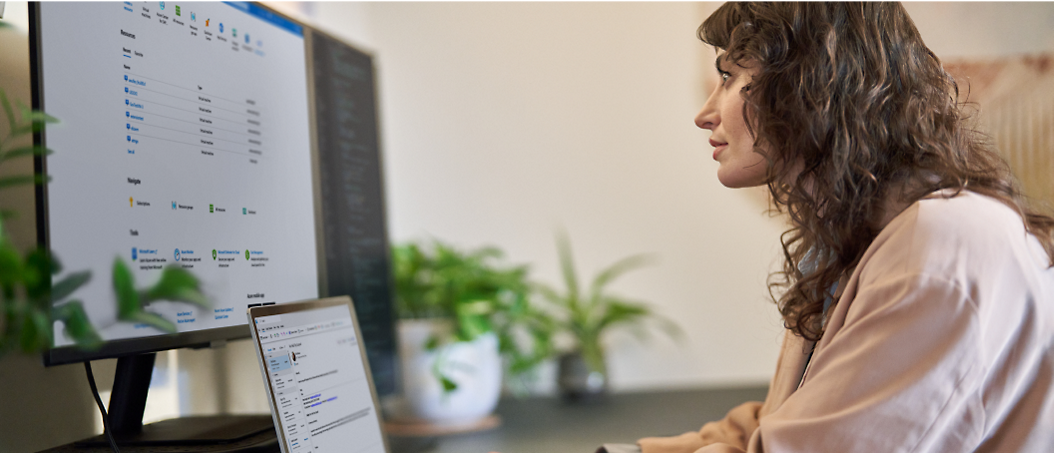 Une femme aux cheveux frisés examine des données sur un écran d’ordinateur dans un bureau, avec des documents et un téléphone visibles.