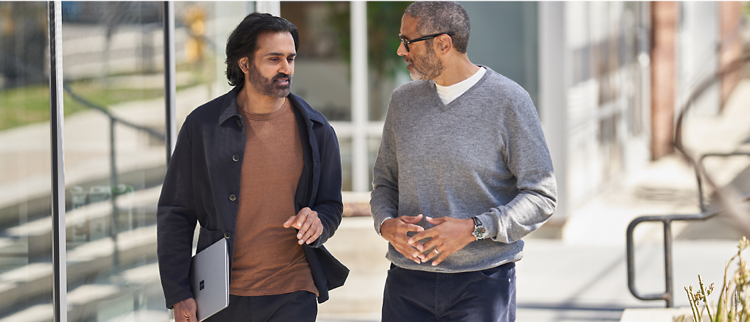 Two men walking and talking on a city sidewalk, one wearing a dark coat and the other in a gray sweater.